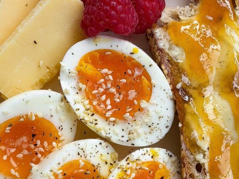 Close-up of soft-boiled egg halves with jammy orange yolks, sprinkled with sesame seeds and black pepper, beside slices of hard cheese, raspberries and seeded toast drizzled with honey.
