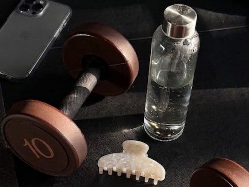 Two dumbbells with bronze plates marked 10 on a dark gym floor, next to an iPhone, a glass water bottle and a hair clip.
