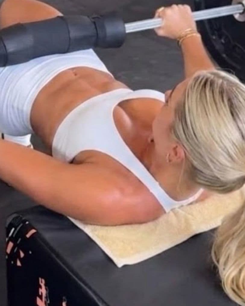 Woman with blonde hair in a white sports bra lying on a gym bench with a towel under her, gripping a loaded barbell above her chest.
