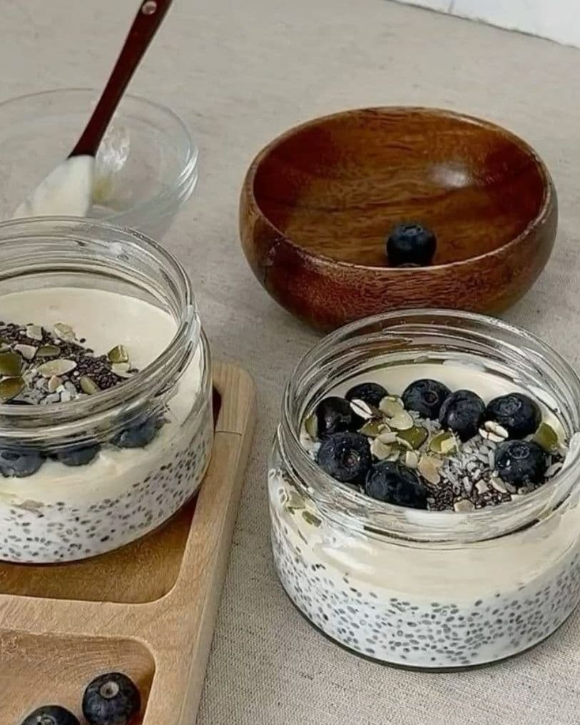 Two glass jars of overnight oats with yogurt, chia seeds, pumpkin seeds and blueberries on a wooden board, next to a wooden bowl and loose blueberries.