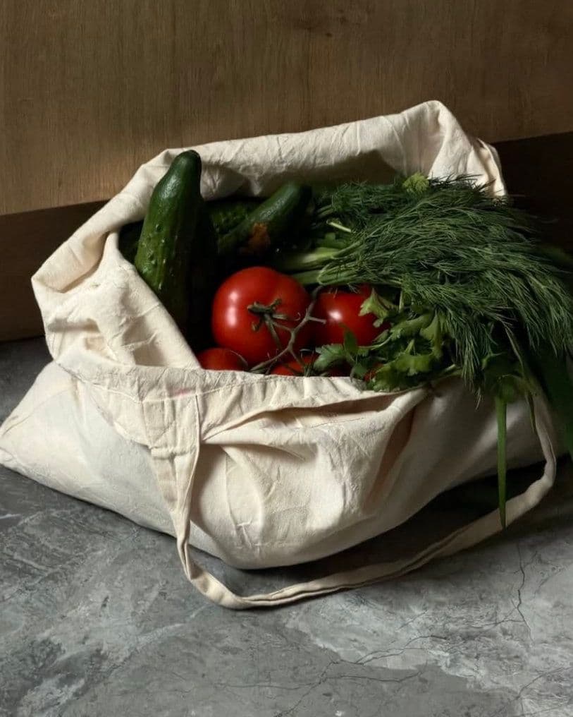 Canvas tote bag on a grey stone surface, filled with tomatoes on the vine, cucumbers and a large bunch of fresh dill.