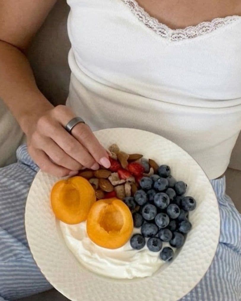 Woman in a white lace-trimmed top holding a plate of plain yogurt topped with halved apricots, blueberries, chopped nuts and a few pieces of strawberry.