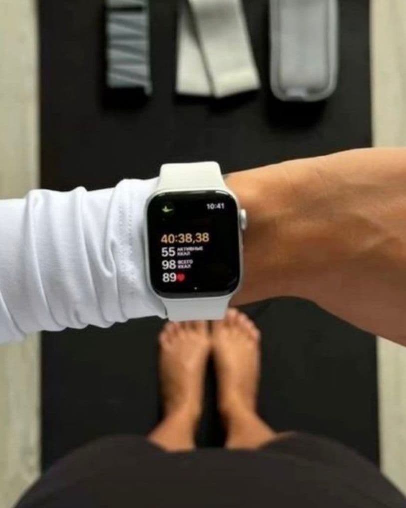 Overhead view of a wrist wearing an Apple Watch with a white band showing a workout timer, above bare feet on a black exercise mat with resistance bands laid out at the top.
