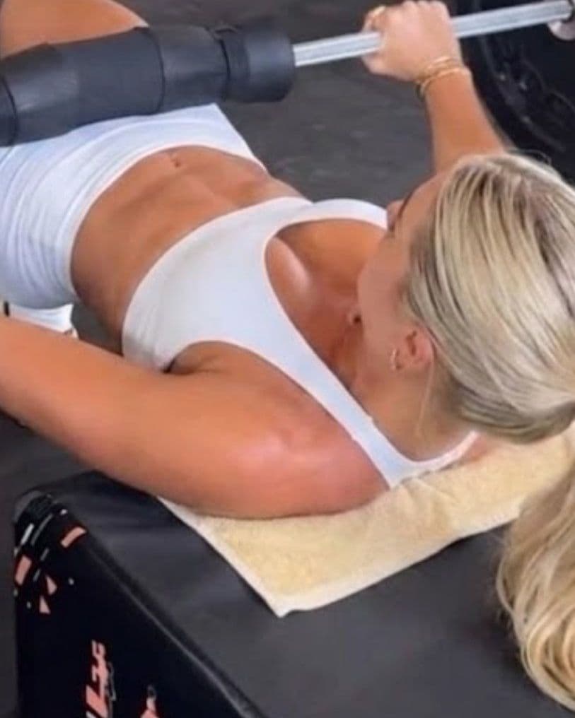 Woman with long blonde hair lying on a gym bench with a towel under her, gripping a barbell for a bench press.