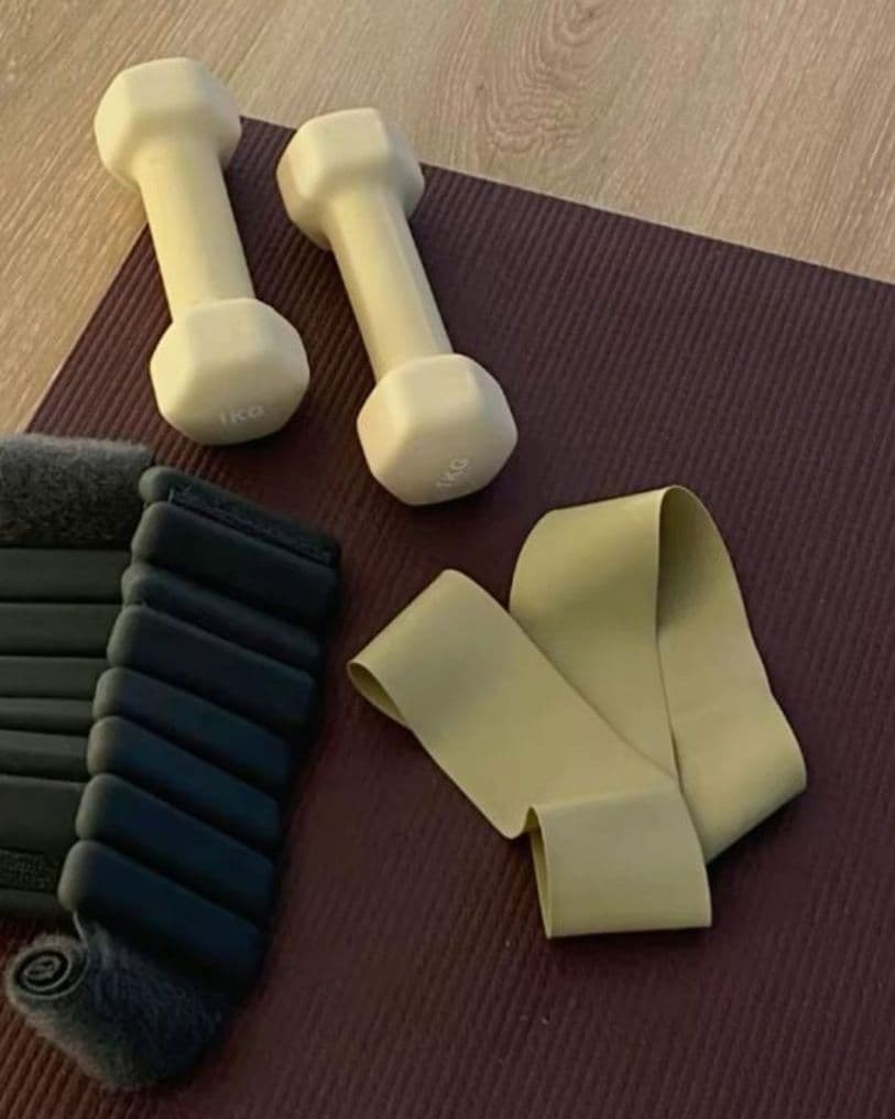 A pair of light cream-colored dumbbells, a looped resistance band and black ankle weights laid on a plum exercise mat on a wooden floor.